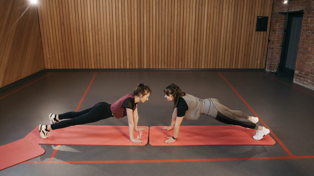 Two women perform planks on red mats in a gym, emphasizing core strength exercise.